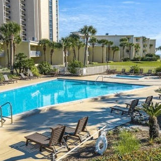 Resort pool and sundeck with lounge chairs, palm trees, and condo towers beside a beachfront lawn.